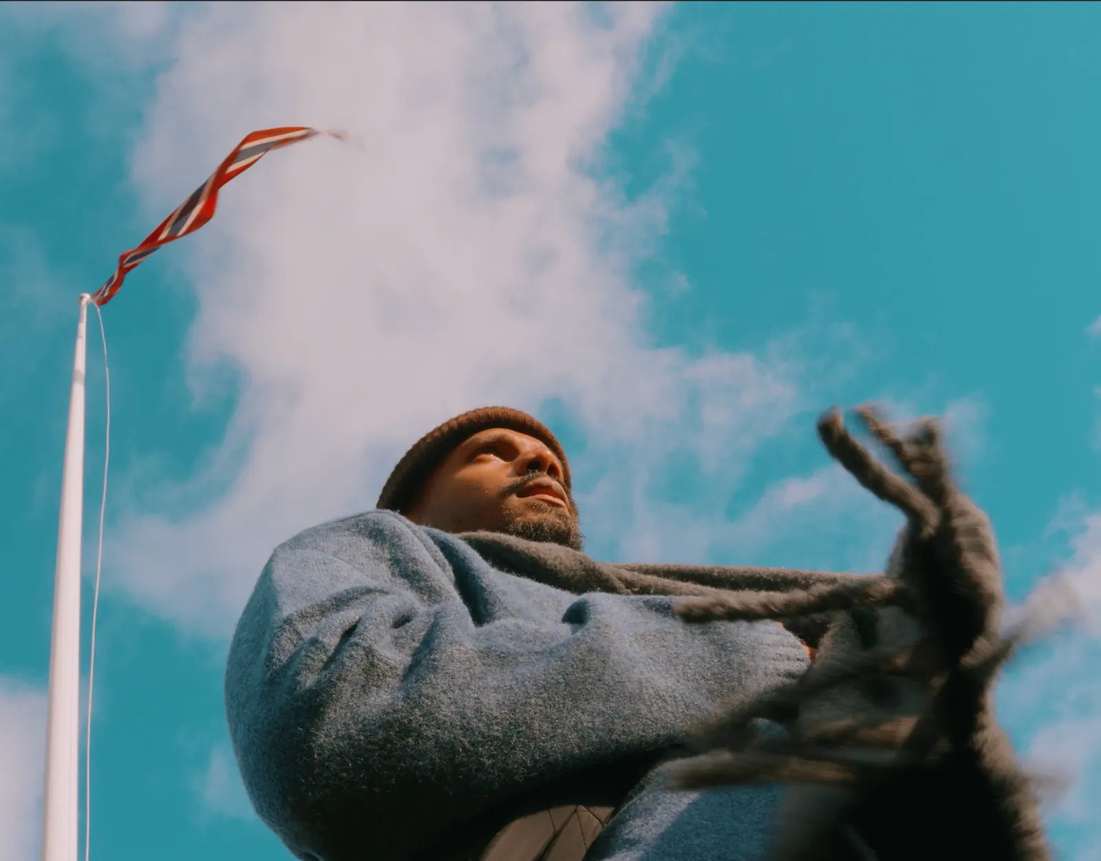 Homme en manteau gris filmé en contre-plongée sous un ciel bleu, un drapeau au vent
