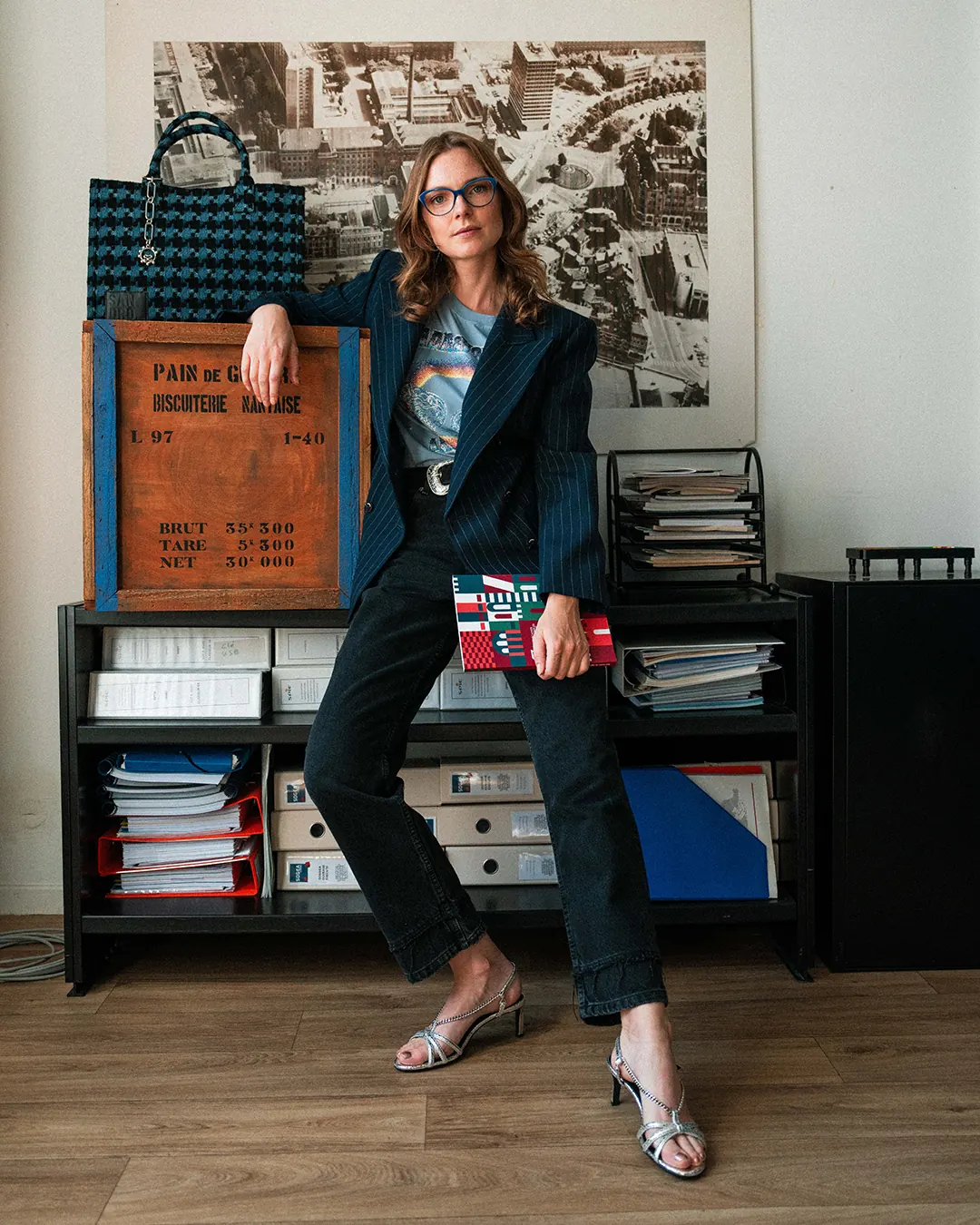 Femme en blazer posant devant un mur d’affiches anciennes, sac à la main
